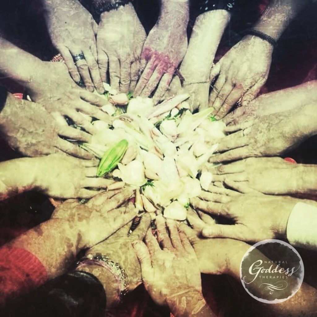 A circle of women's hands touching a bowl of flowers.